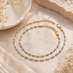 A single-strand gold and silver chain anklet with details, arranged in a loop on a beige textured fabric with dried floral props.